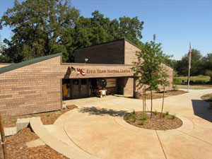 Exterior of the Effie Yeaw Nature Center, a brown-brick building with a sign and open doors, with a flag pole and trees on a sunny day.