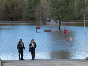 Two park rangers stand on a flooded road, observing a flooded park. Water surrounds trees, signposts, and a building in the background. A stop sign and other park signs are visible amidst the high water.