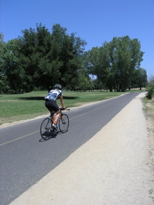 Cyclist riding a road bike on a paved path next to a sandy shoulder, with trees and grass in a sunny park setting.