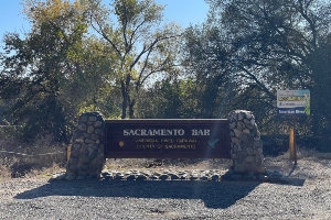 Large wooden sign at Sacramento Bar, a historic gold rush site, with "SACRAMENTO BAR" in large letters, text below indicating it is a part of the American River and part of the City and County of Sacramento. The sign is flanked by stone pillars and set against a backdrop of trees and a clear blue sky.