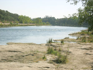 A wide river scene with a rocky, sandy foreground, calm blue water, and tree-lined banks under a hazy sky.