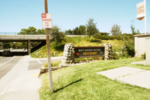 Sign indicating "Watt Avenue Access" with a stone-bordered entrance, under a bridge and a "No Parking" sign in the foreground, with trees and a light blue sky in the background.